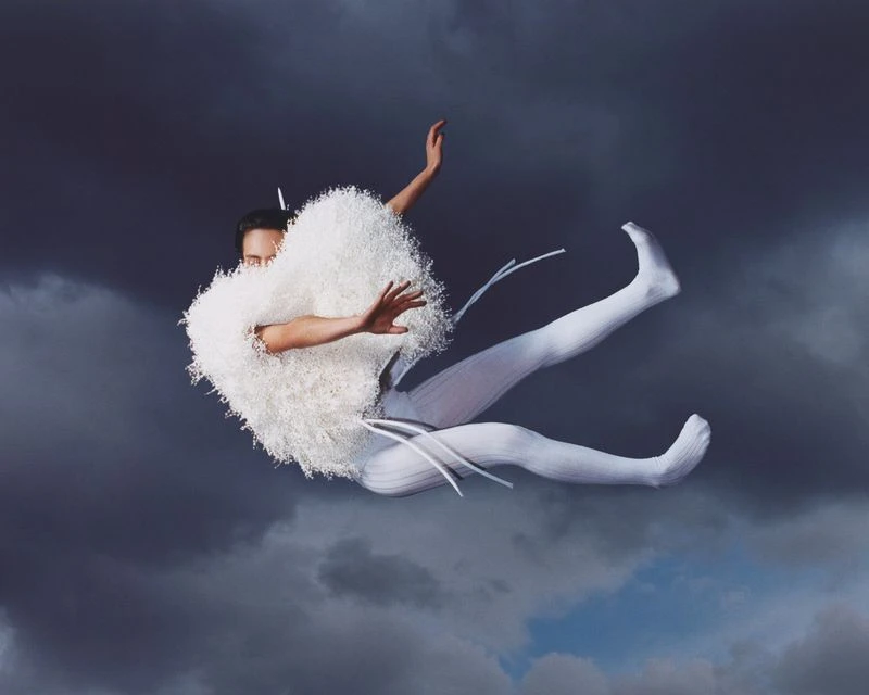Person in white costume levitating against a stormy sky. The outfit features fluffy shoulders and textured leggings, creating an ethereal effect.