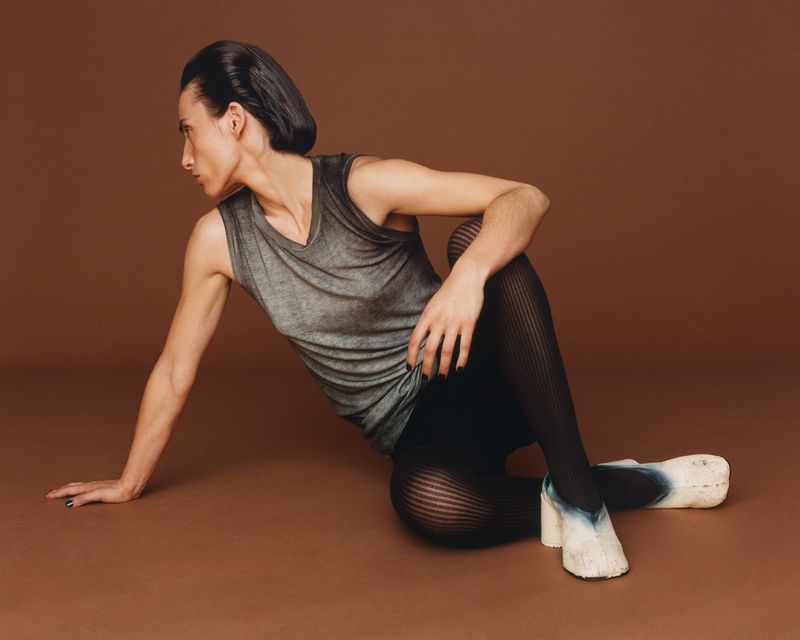 Woman in gray tank top and black tights poses on a brown seamless background looking left, seated with legs crossed and wearing mule shoes.