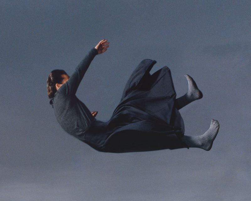 A woman in mid-air wearing a long, dark dress and socks against a muted, grayish-blue sky; the photo gives the impression of floating.