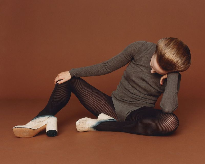 Woman in gray ribbed top and patterned tights seated against a brown backdrop, paired with chunky platform shoes, studio shot.