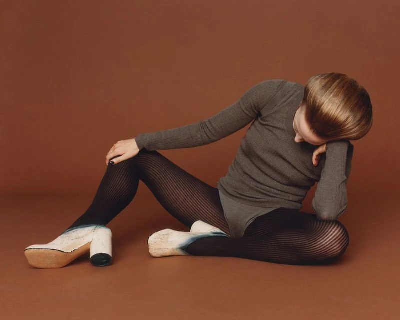 Woman in gray ribbed top and patterned tights seated against a brown backdrop, paired with chunky platform shoes, studio shot.