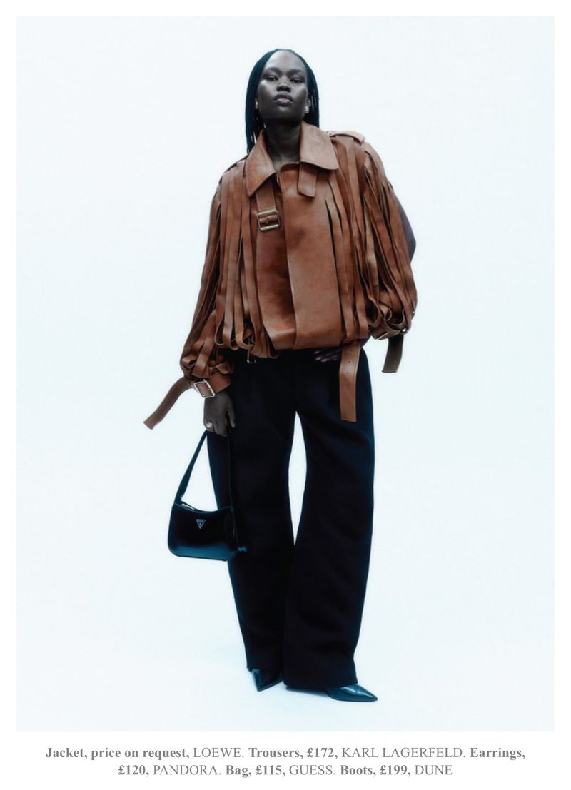 Fashion editorial shot of a Black female model posing in a brown jacket, black trousers, a black purse, and black heels against white backdrop.