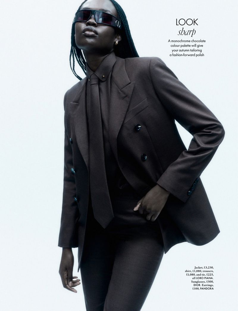 Dark-skinned woman poses in a chocolate-brown pantsuit with sunglasses and a tie against a bright background in a fashion editorial shot.