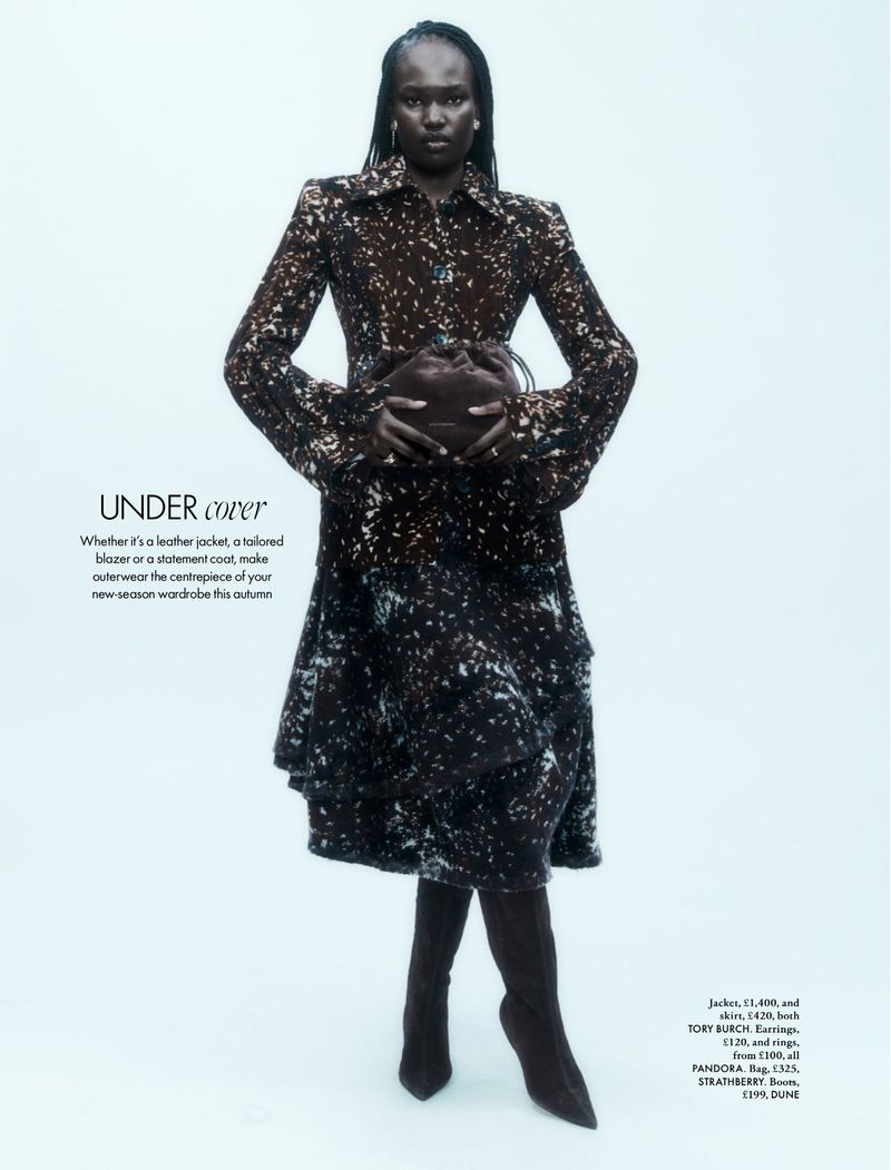 Fashion editorial shot featuring a black woman in a patterned coat dress and boots holding a small handbag against a white backdrop.