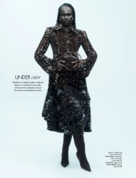 Model Adual Akol posing in a speckled brown jacket, matching skirt, and pointed boots against a pale blue studio backdrop.