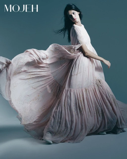 A fashion shot of a woman in a flowing pink gown with scattered gold flecks against a neutral studio background, creating a soft, dreamy aesthetic.