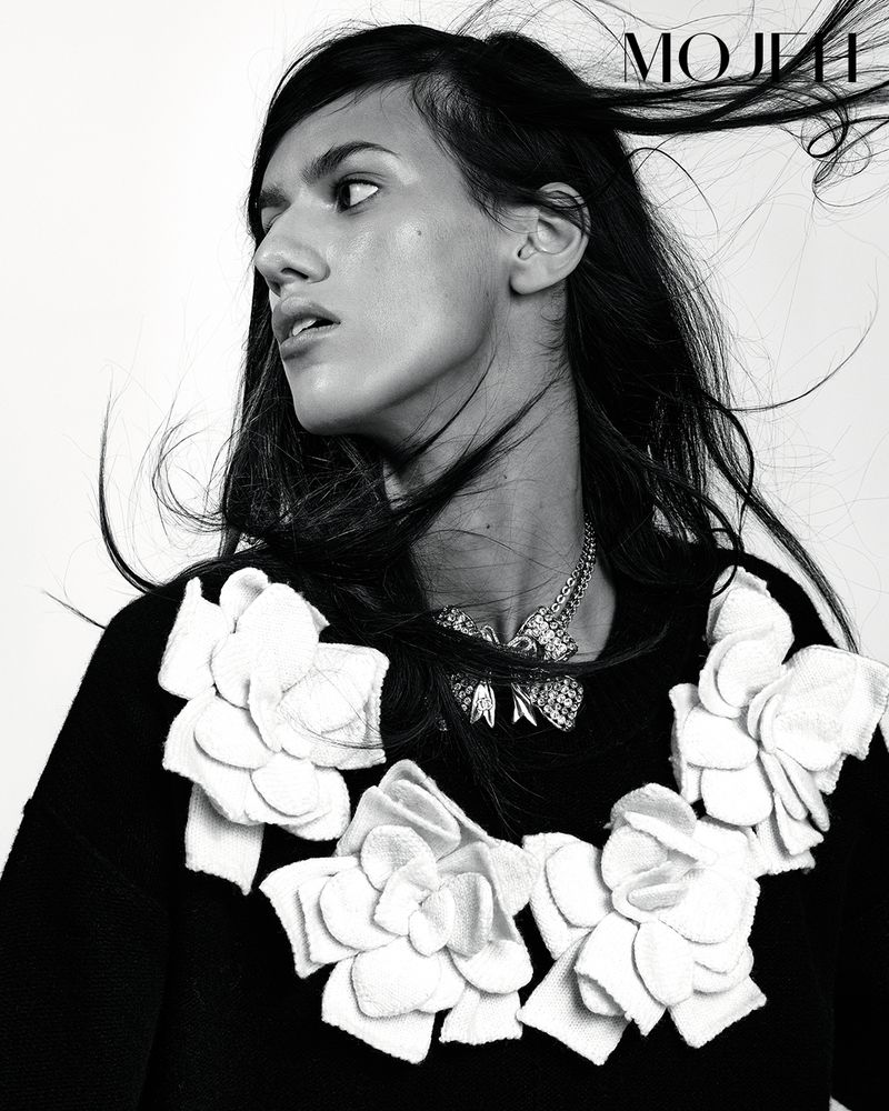 Monochromatic fashion portrait of a young woman with dark hair, statement necklace, and black top with white flower embellishments.