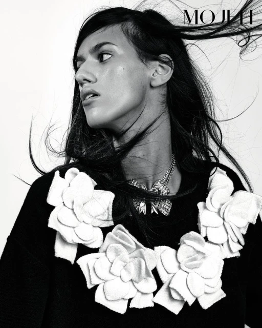 Monochromatic fashion portrait of a young woman with dark hair, statement necklace, and black top with white flower embellishments.