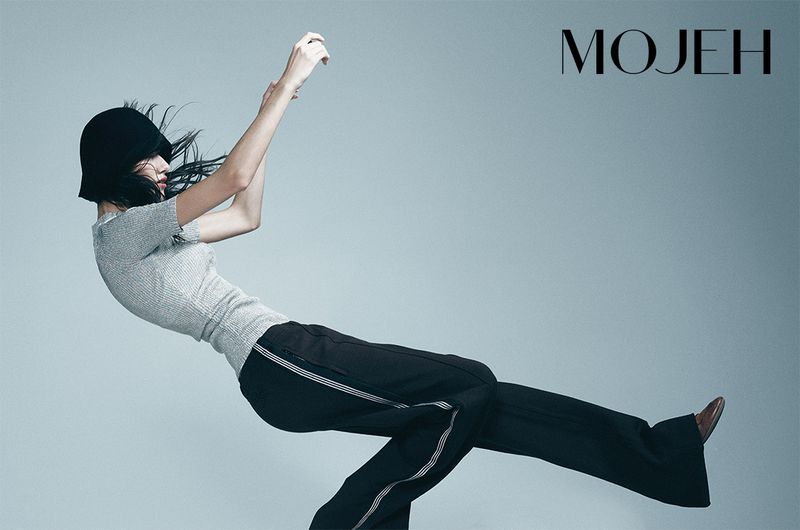 Fashion editorial shot of a slender adult woman wearing a black hat, grey top and black pants, posing in a studio with a light-blue background.