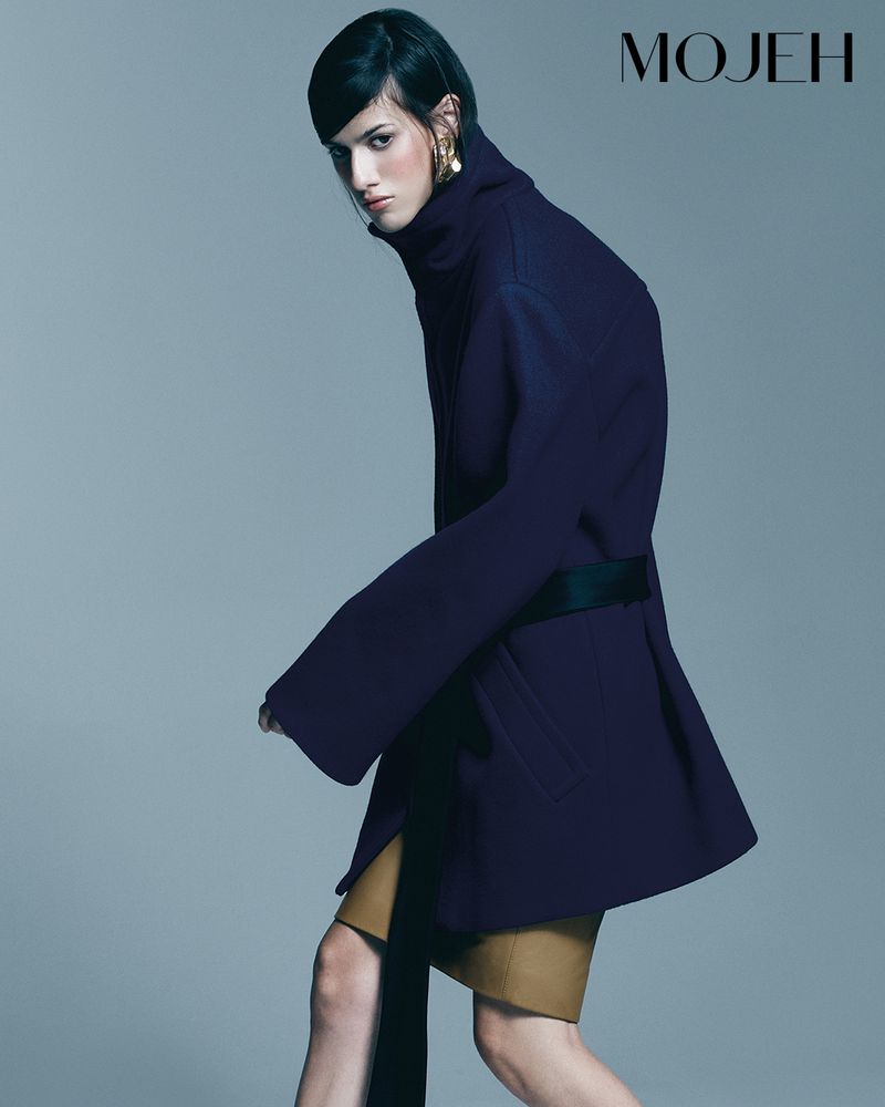 Elegant adult woman in a studio fashion shot wearing a stylish blue belted jacket and a tan skirt, posing with confidence in a grey studio.