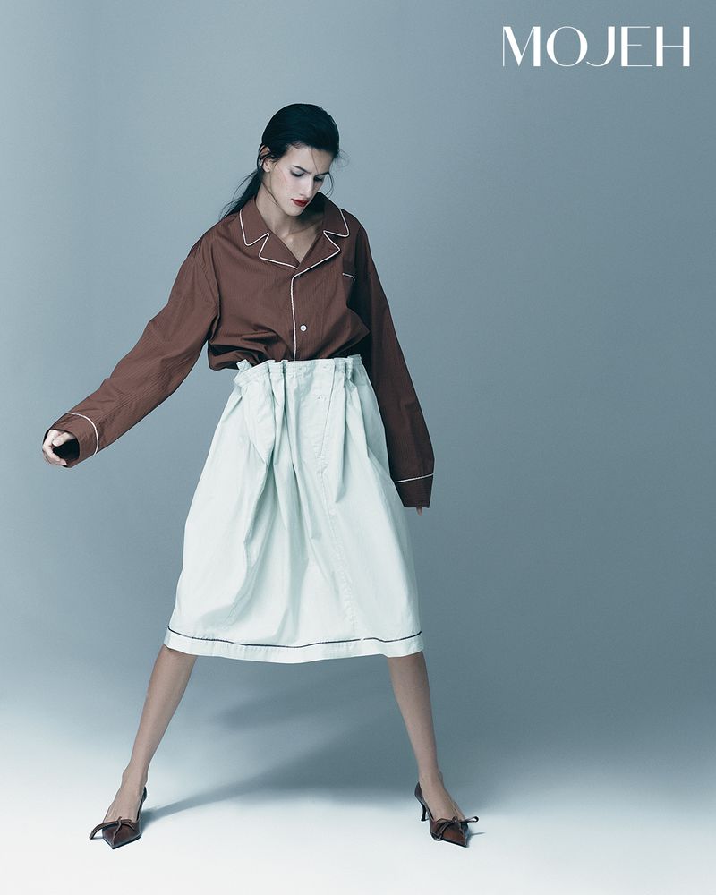 Fashion editorial shot of a female model in a brown long sleeve blouse and pale green skirt, posing in a studio with soft lighting.