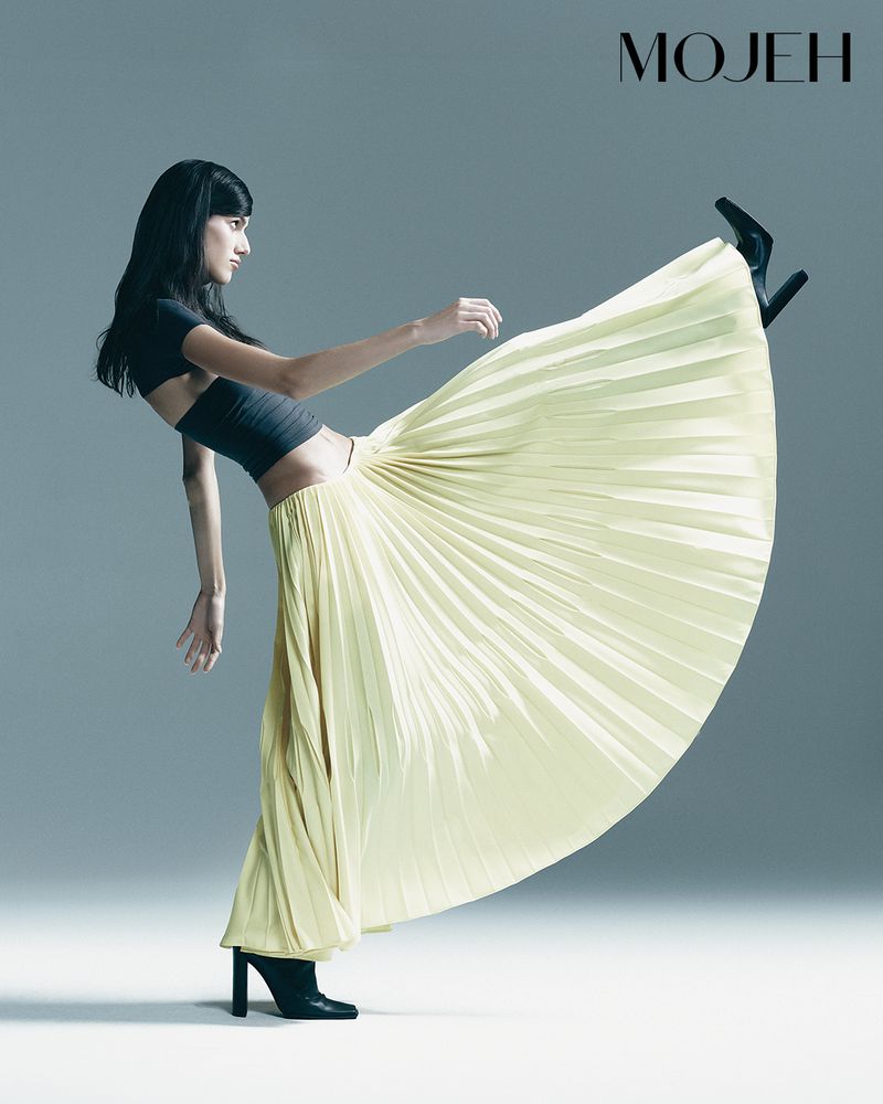 Fashion shot of a woman in a pleated yellow maxi skirt, black crop top, and black heeled boots striking a dynamic pose against a gray backdrop.