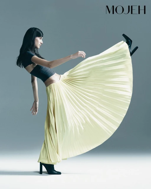 Fashion shot of a woman in a pleated yellow maxi skirt, black crop top, and black heeled boots striking a dynamic pose against a gray backdrop.