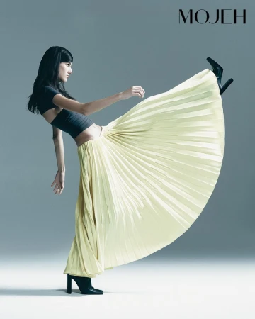 Fashion shot of a woman in a pleated yellow maxi skirt, black crop top, and black heeled boots striking a dynamic pose against a gray backdrop.