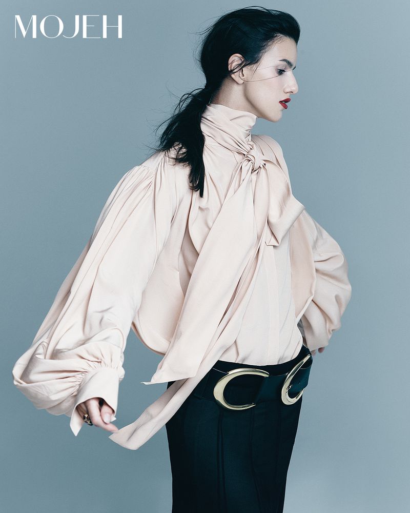 Fashion editorial shot of a woman in a stylish cream-colored blouse with a large bow, black skirt, and a statement belt on a grey background.