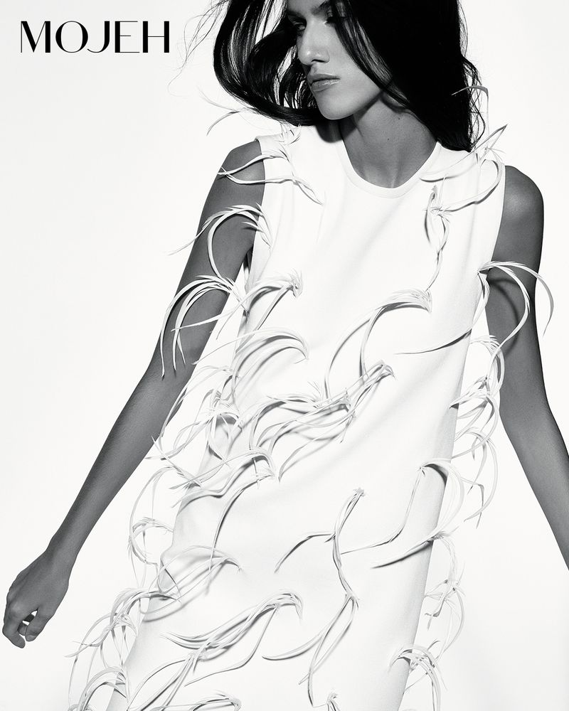 Model wearing a white sleeveless dress with sculptural feather-like fringe details against a white background in black and white.