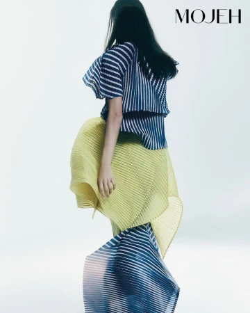 Fashion shot of a woman wearing a flowing striped top and a pleated yellow skirt, posing in a bright studio with dramatic movement.