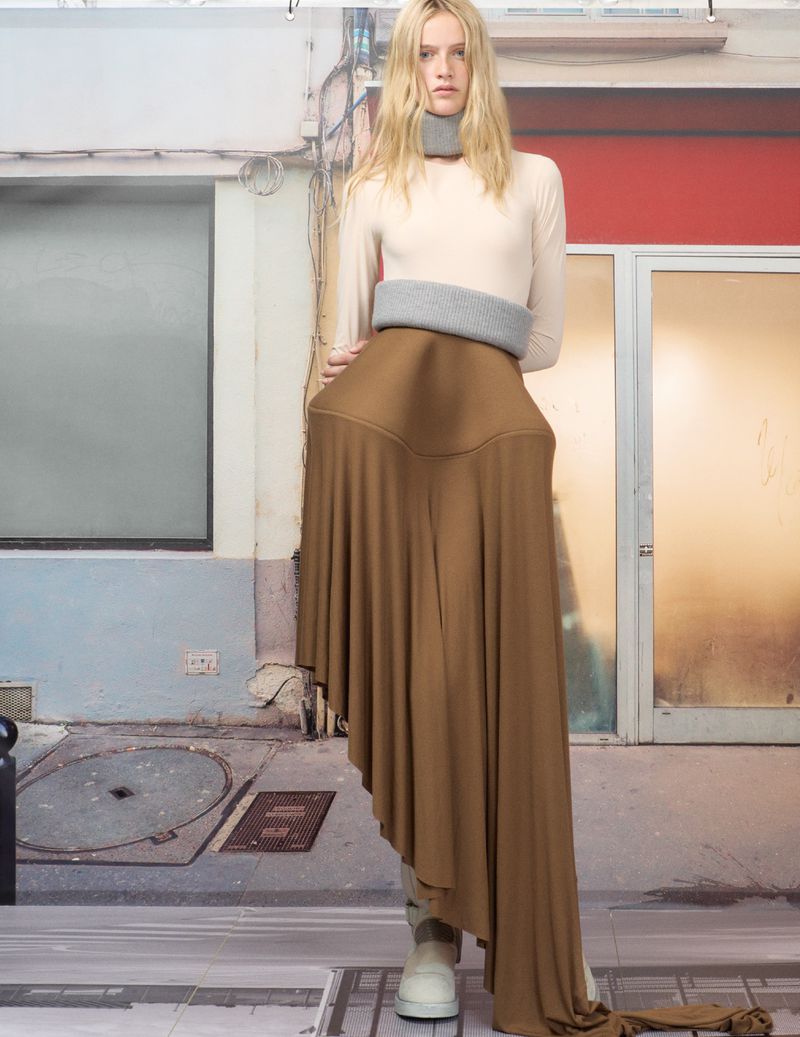 A young woman models a draped, asymmetrical, neutral-toned skirt, a top, and gray knit neckwear against a digitally imposed urban backdrop.