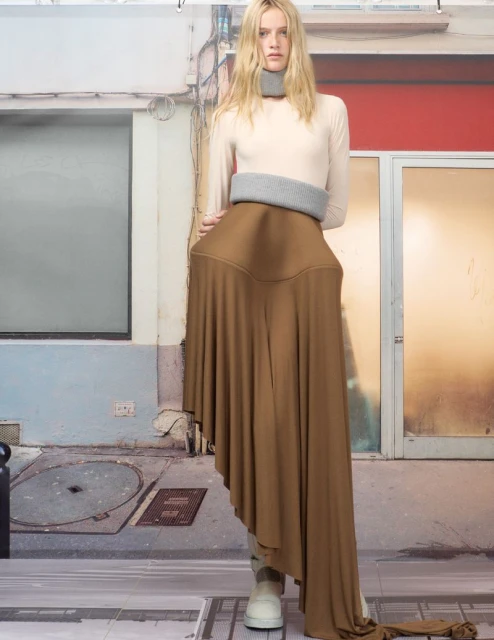 A young woman models a draped, asymmetrical, neutral-toned skirt, a top, and gray knit neckwear against a digitally imposed urban backdrop.