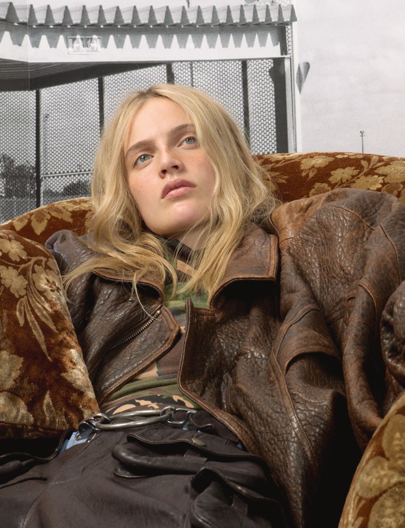 Young woman poses on an ornate couch wearing a brown leather jacket and dark skirt in an indoor setting with a patterned backdrop.