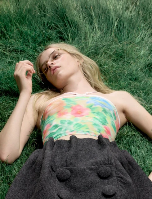 Young woman with blonde hair and glasses lying in green grass wearing a floral top and gray skirt in a natural outdoor setting.