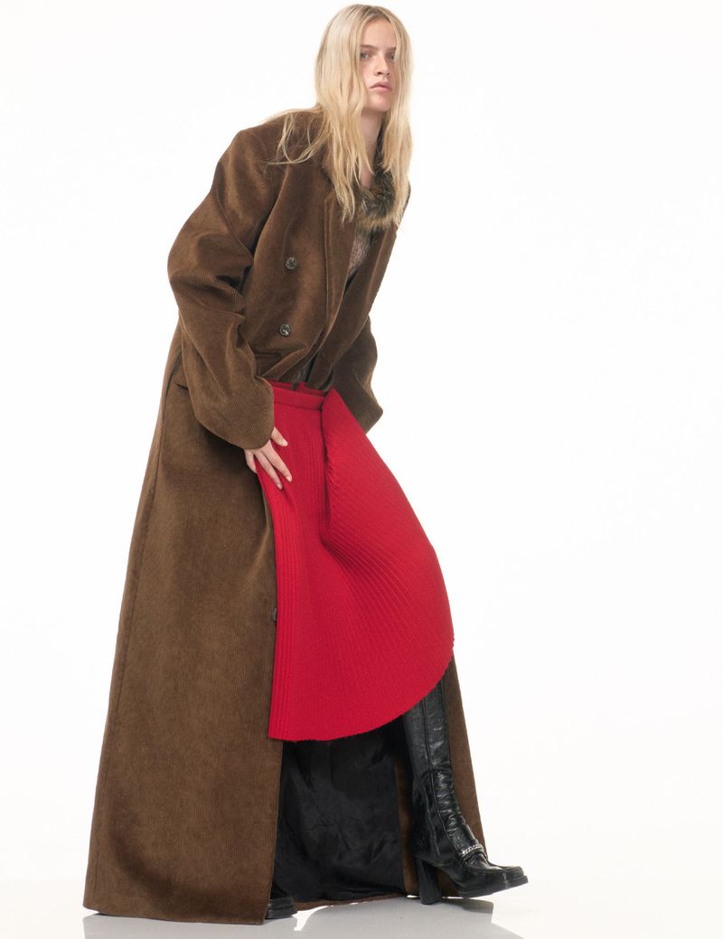 Fashion shot of a blonde woman wearing a brown, long overcoat with a red skirt, and black boots, posing in a photo studio.
