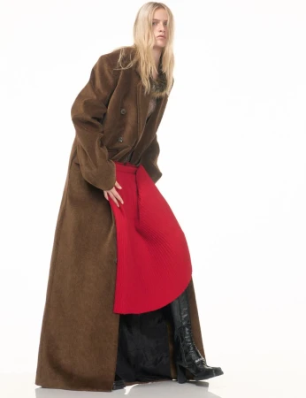 Fashion shot of a blonde woman wearing a brown, long overcoat with a red skirt, and black boots, posing in a photo studio.