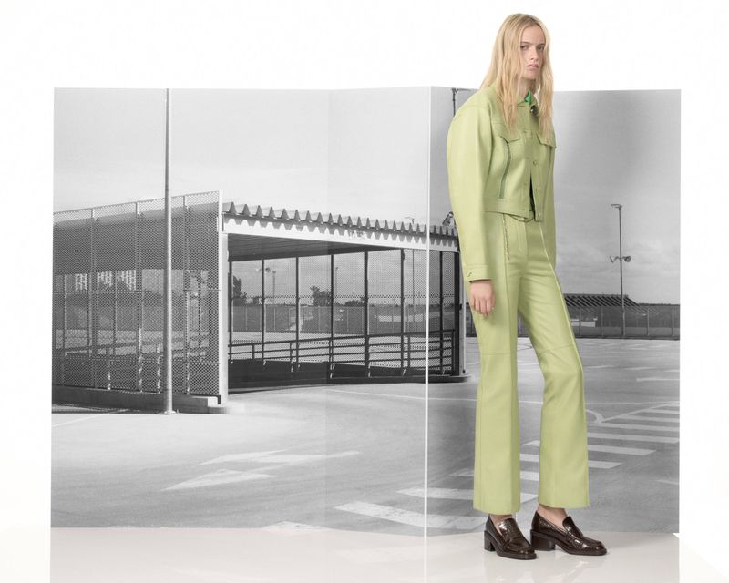 A slender woman poses in a pistachio-green suit in front of a black and white urban background, displaying fashion and style.