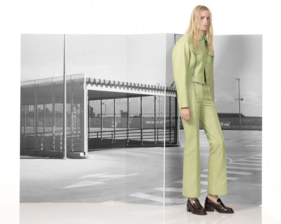 A slender woman poses in a pistachio-green suit in front of a black and white urban background, displaying fashion and style.