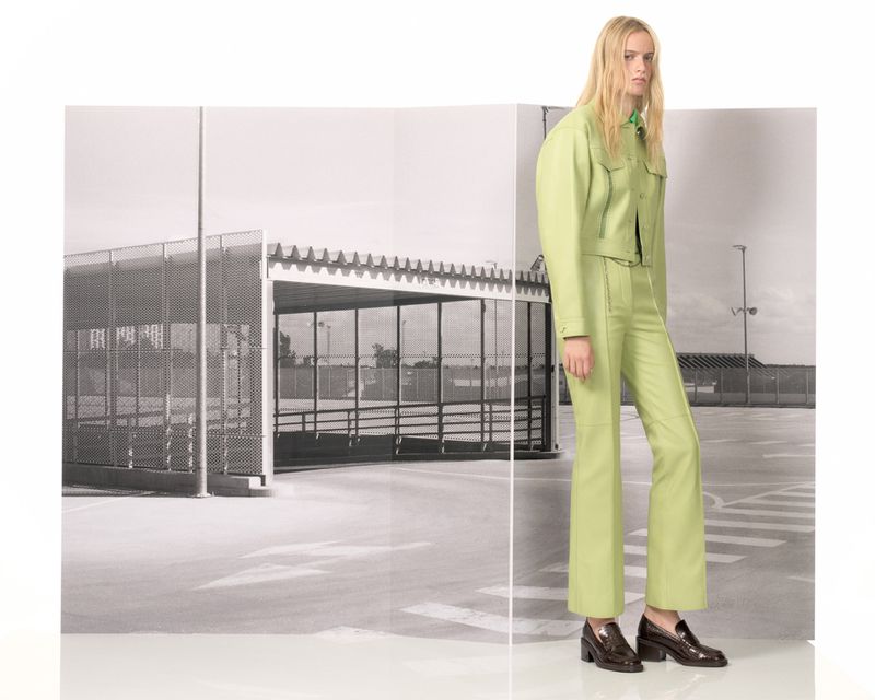 Fashion shot of a woman in a light green jacket and pants, standing against a monochrome background of a bus stop structure.