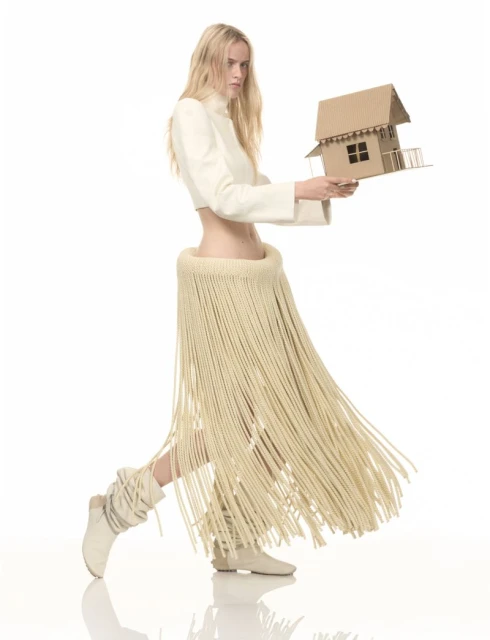 Fashion model poses in a white cropped jacket and a long fringe skirt holding a cardboard model house on a white background.