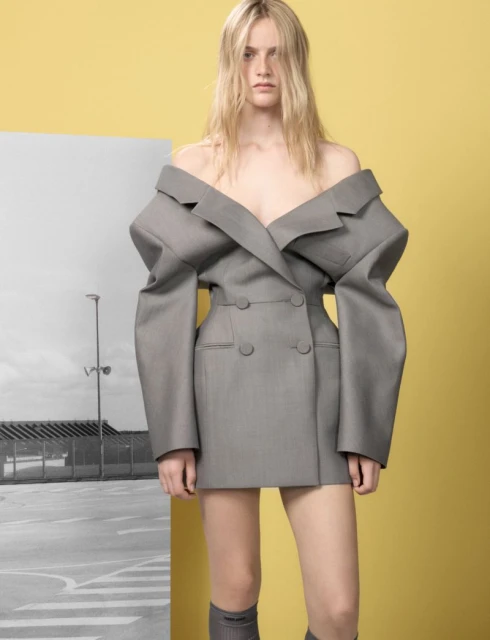 Fashion shot of a blonde adult female model wearing an off-the-shoulder gray blazer dress against a yellow background with a grayscale backdrop.