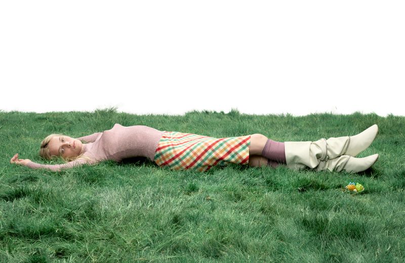 Model Lauren Huyskens lying horizontally across green grass wearing a pink knit top, plaid skirt, and slouchy cream boots.