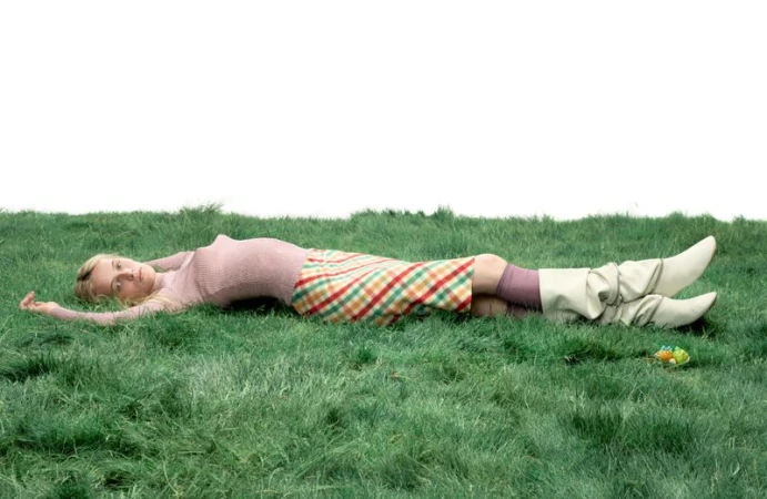Model Lauren Huyskens lying horizontally across green grass wearing a pink knit top, plaid skirt, and slouchy cream boots.