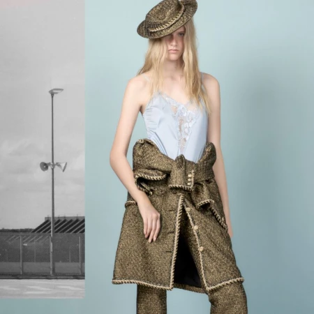 Fashion shot of a blonde model wearing a light blue top, gold and black patterned jacket and hat, against a split studio background.