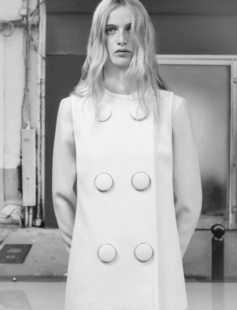 Blonde woman in a white dress with large buttons stands in front of a textured wall in a black and white fashion photograph.