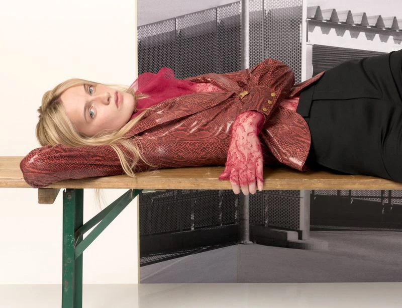 Blonde woman lying on a wooden table, wearing a red snakeskin jacket and black pants, with a neutral expression, and a monochrome background.