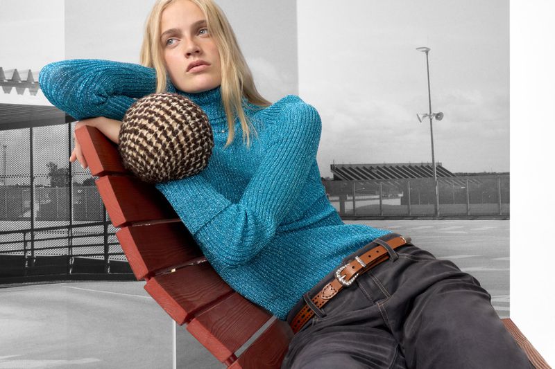 A fair-skinned female model reclines in a turquoise knit sweater, gray jeans and brown belt on a red bench with a brown checkered sphere.