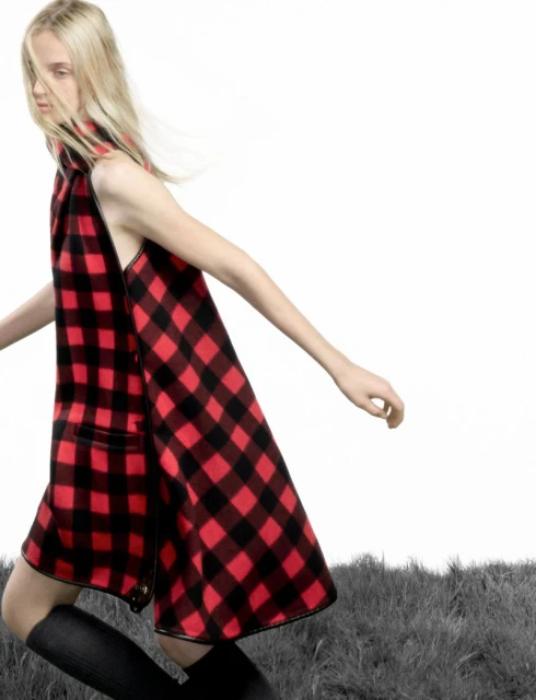 Fashion shoot of a blonde woman wearing a red and black checkered dress. She is walking in a field with a blurred background.