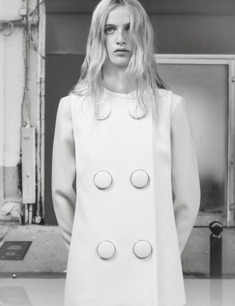 Fashion portrait of an adult woman with long blonde hair wearing a white dress with buttons against a gray industrial background.