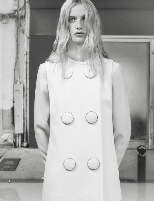 Fashion portrait of an adult woman with long blonde hair wearing a white dress with buttons against a gray industrial background.