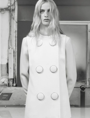Fashion portrait of an adult woman with long blonde hair wearing a white dress with buttons against a gray industrial background.