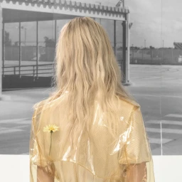 Back view of a blonde woman wearing a translucent yellow vinyl coat with a single yellow daisy tucked into the back.