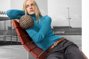A blonde model leans back on a red wooden slatted lounger wearing a vibrant turquoise ribbed turtleneck sweater and dark trousers.