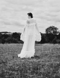 Model wearing a sheer floor-length white lace gown standing in an open field under a cloudy sky in black and white.