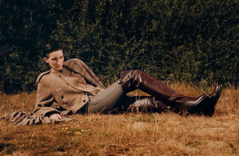 Model lying on dry grass wearing a brown wool jacket and tall leather boots outdoors