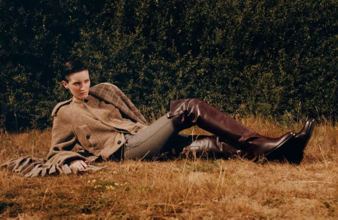 Model lying on dry grass wearing a brown wool jacket and tall leather boots outdoors