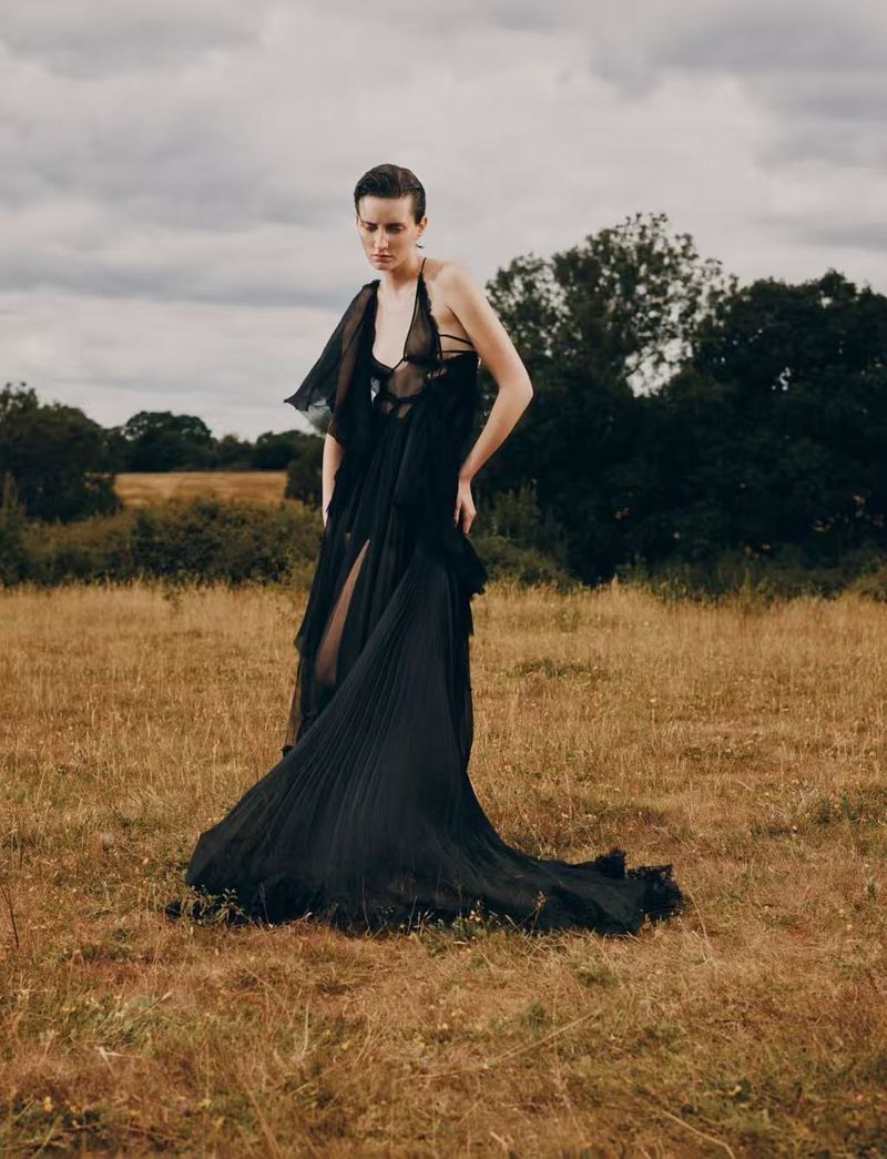 Woman in a flowing black gown standing in a golden field with trees and cloudy sky in the background, fashion editorial photography.