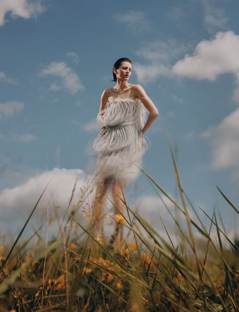 A woman in a fringed dress poses in a field of wildflowers under a bright sky with clouds, creating a surreal, dreamy atmosphere.