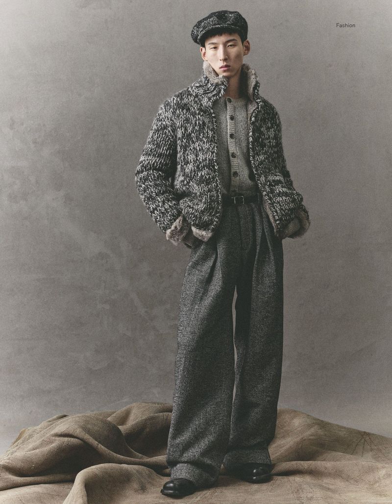 A fashionable Asian man models a tweed driving cap, cardigan sweater, grey suit pants, and black loafers against a grey backdrop.